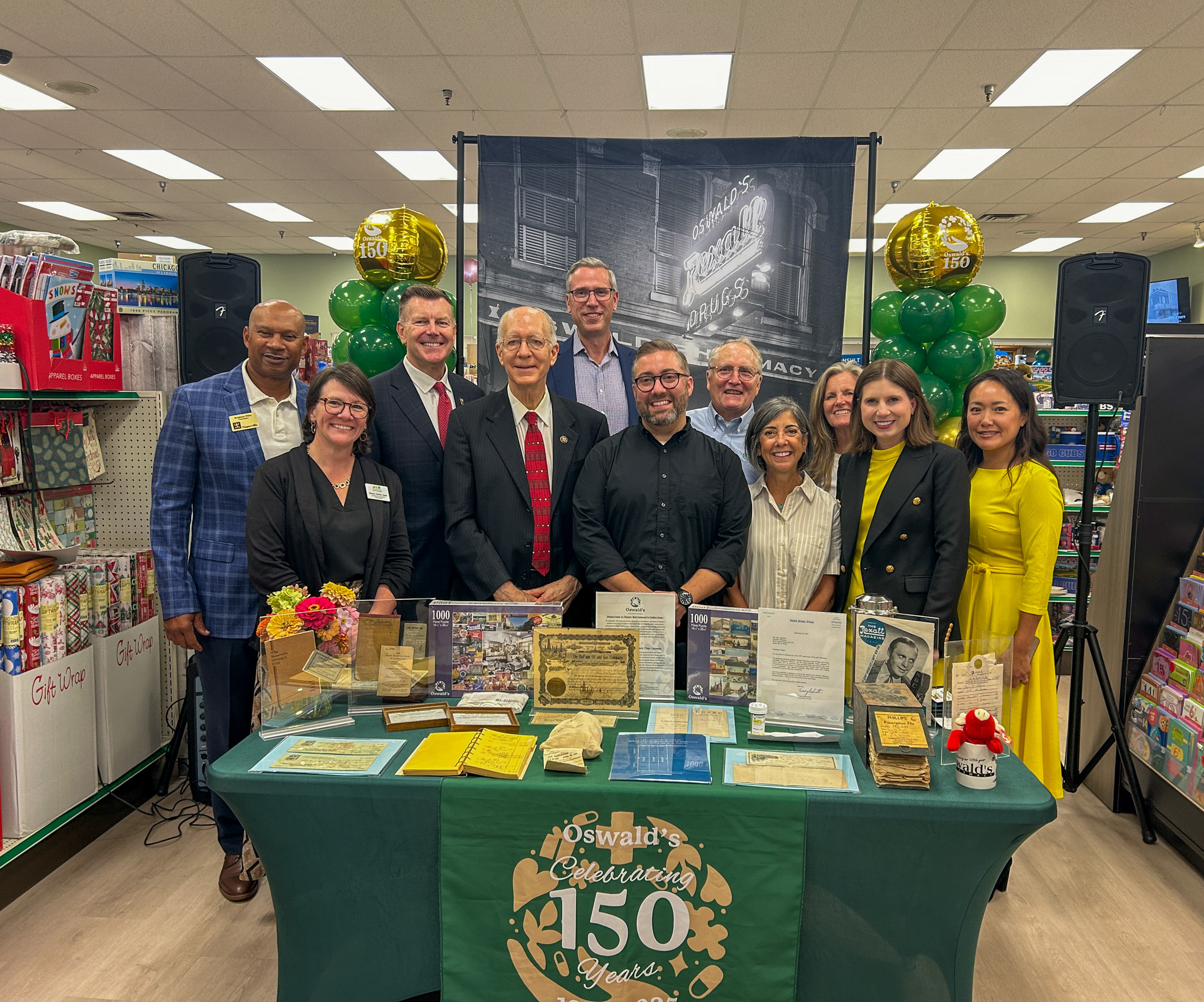 Treasurer Frerichs stands with about 10 pharmacy employees behind a table with a cloth bearing a message about Oswald Pharmacy's 150th anniversary