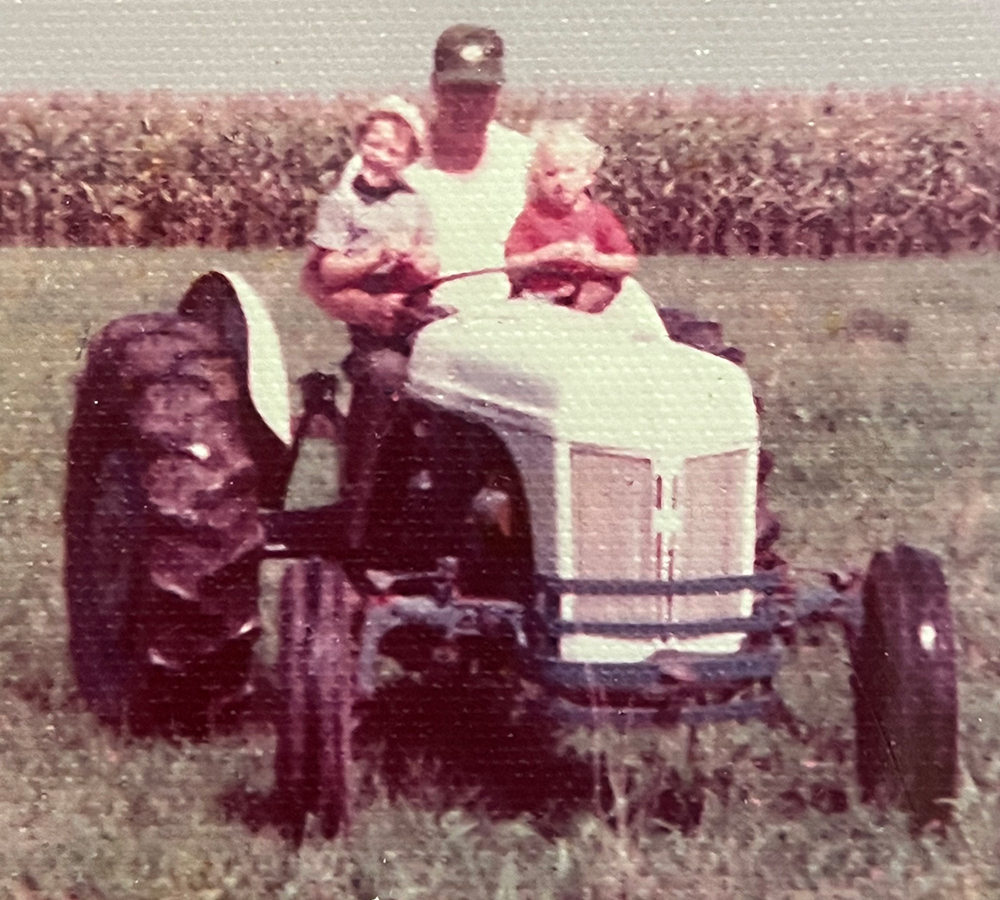 A man sits on a tractor in the field. He has two small boys in his lap.