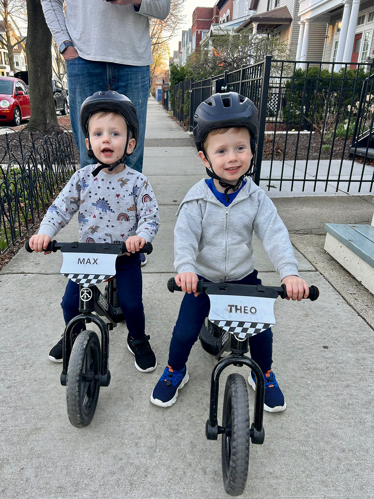 Twin toddler boys riding small bikes on a sidewalk outside