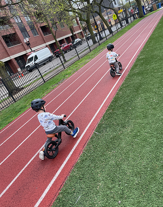Twin toddlers riding small bikes outside on a track and wearing helmets